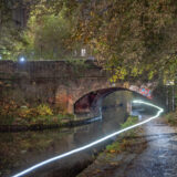 An image of the Ashton Canal in Manchester at dusk. There is an old bridge, and from under it a cyclist has emerged, leaving a trail of white light.